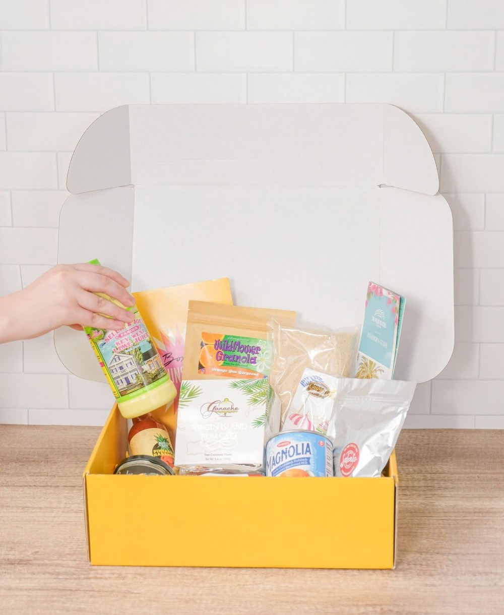 Hand placing a bottle in a yellow gift box with assorted items on a wooden table.