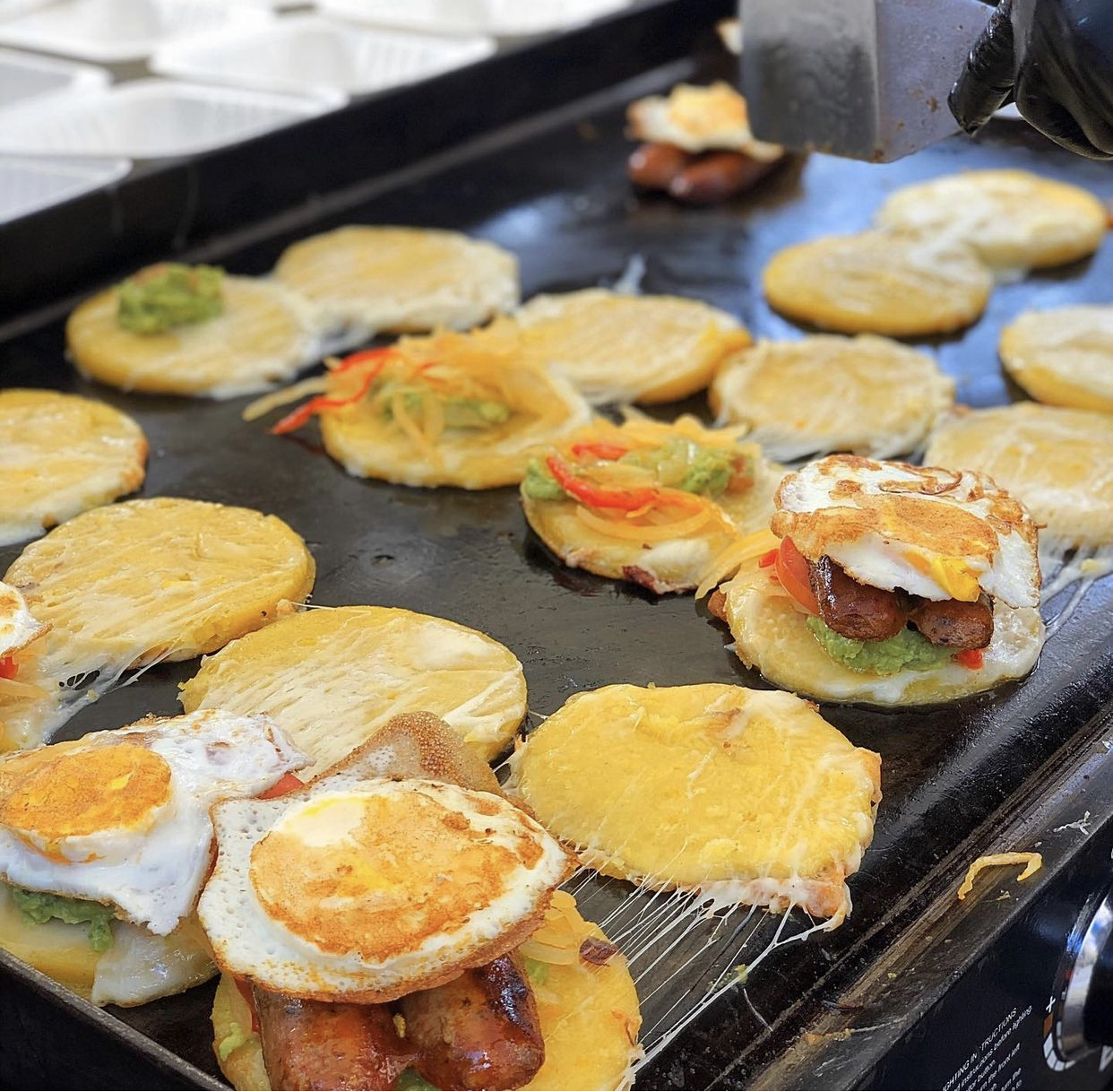 Griddle with cheese arepas, toppings, and fried eggs cooking.