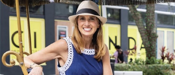 Smiling woman in a hat and blue dress sitting outdoors beside a wooden railing.