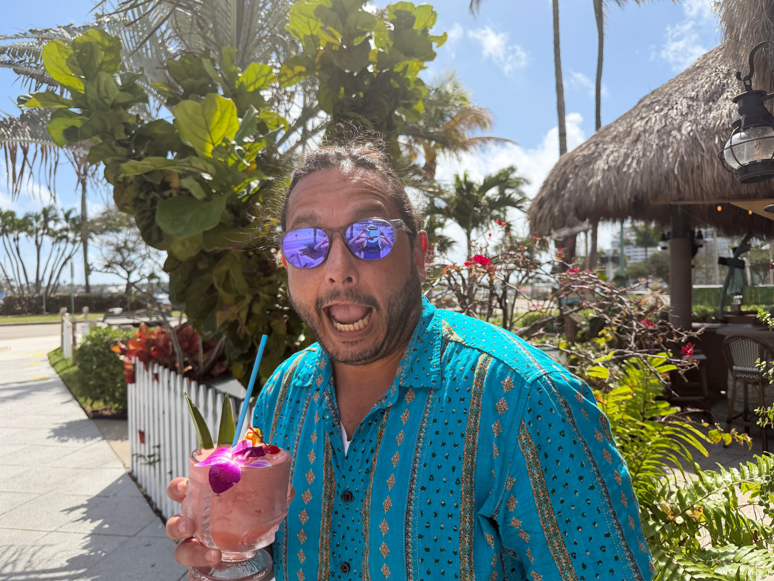 Man in blue shirt holds a drink with a purple flower, standing outdoors near tropical plants.