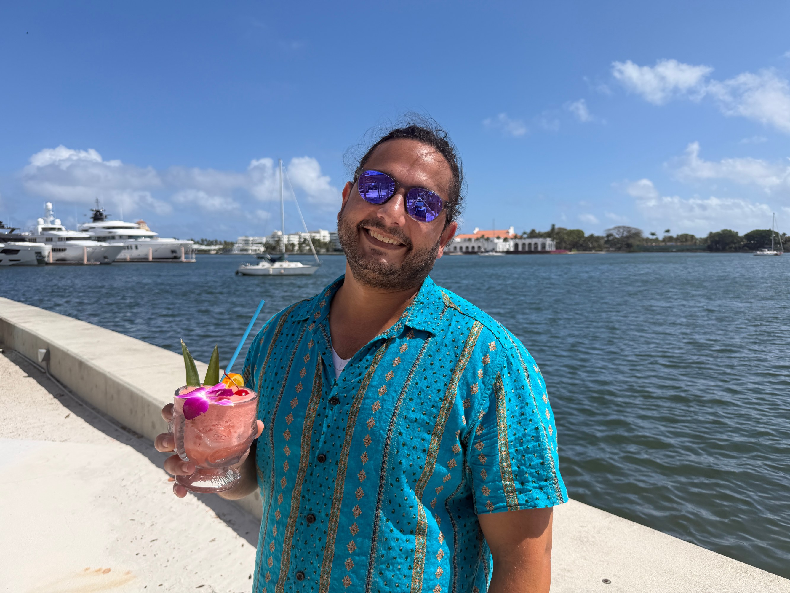 Man in blue shirt holding a tropical drink by the water with yachts in the background.