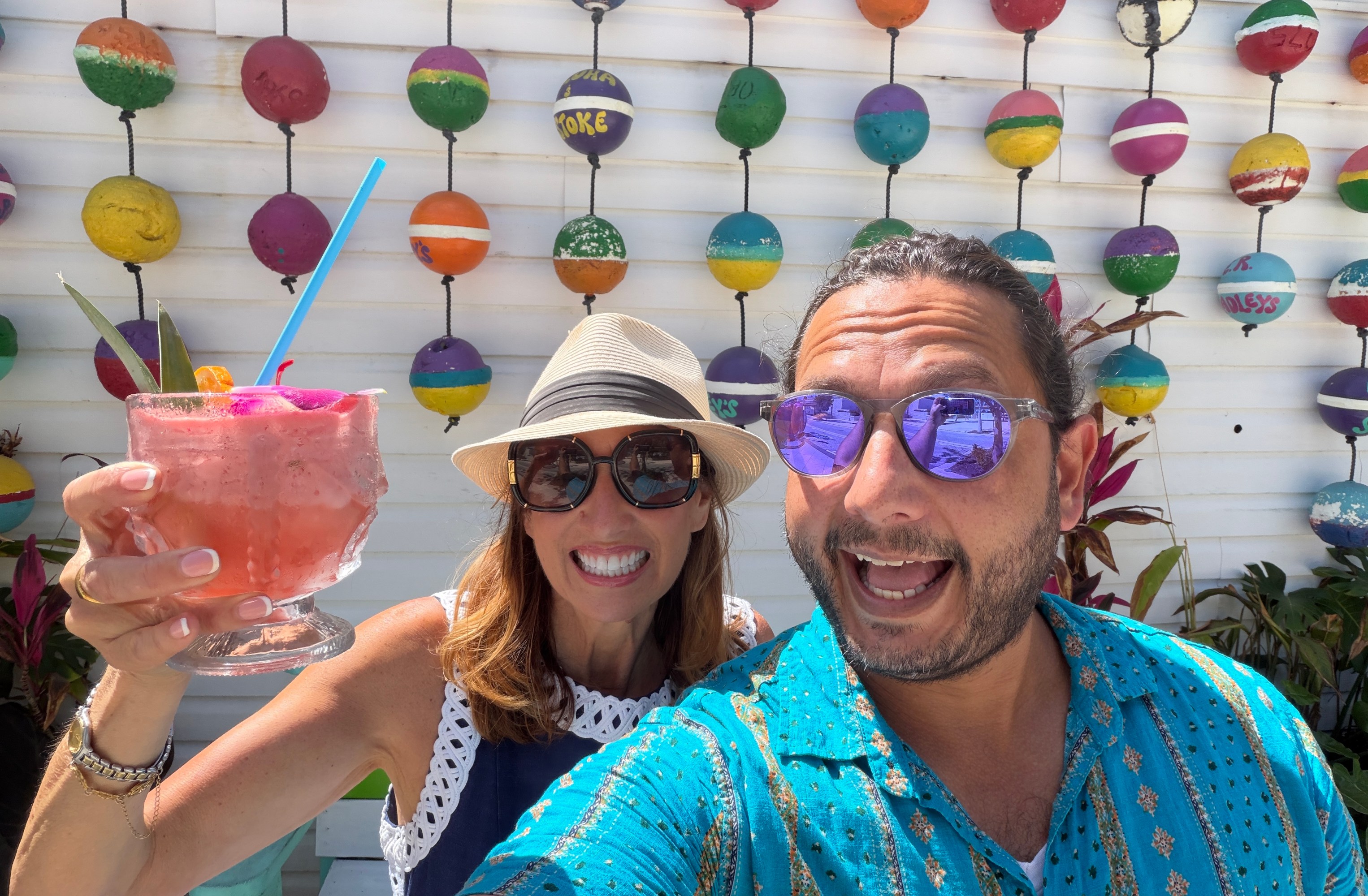 Two smiling people with a tropical drink, colorful buoys hanging in the background.