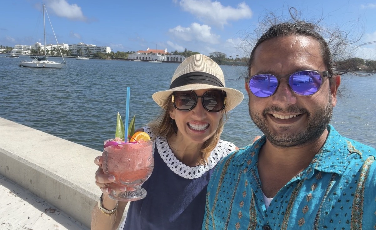 Smiling couple with drinks by the water, wearing sunglasses and hats, with a sailboat in the background.