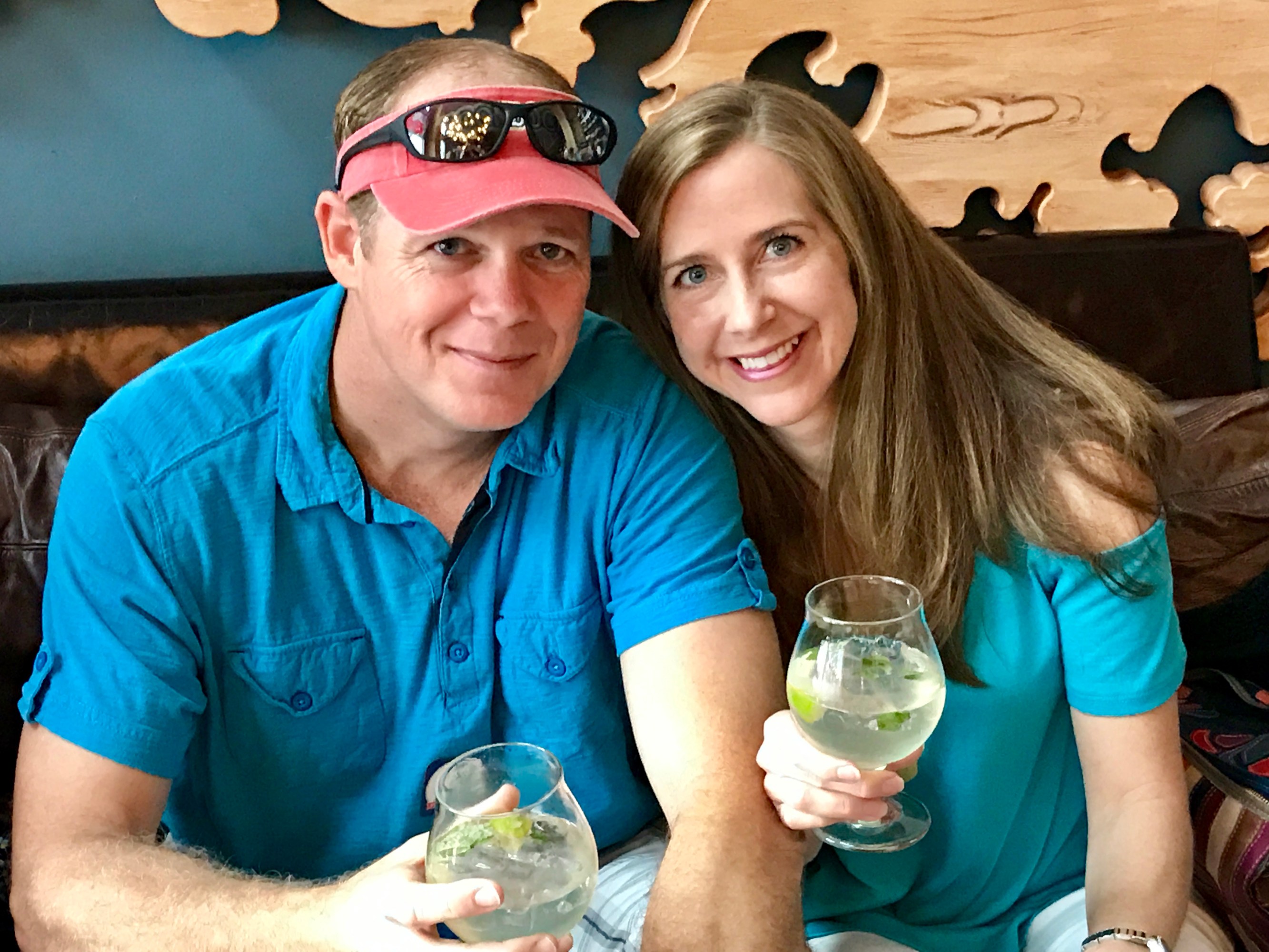 Man and woman in blue tops holding drinks, sitting on a couch, smiling.